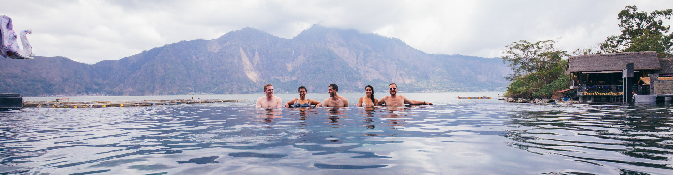 Group talking and laughing in infinity pool in front of mountain on a cloudy day in Bali