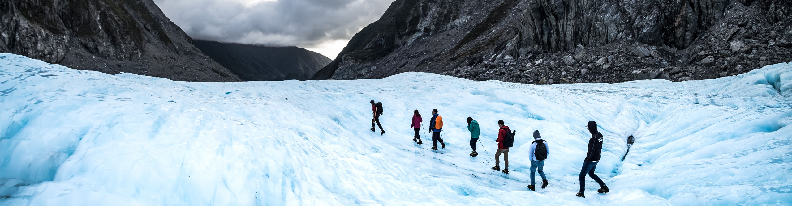 Group hiking across the ice at Fox Glacier in between the mountains on a cloudy day, New Zealand