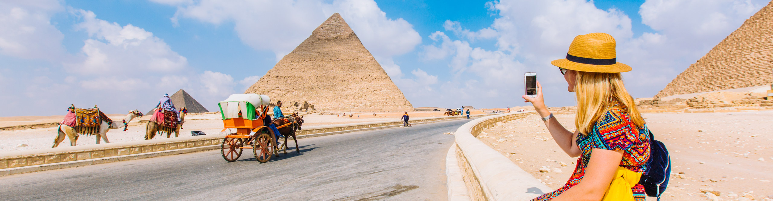 Woman taking pictures of pyramid with people riding camels nearby on a sunny day, Egypt