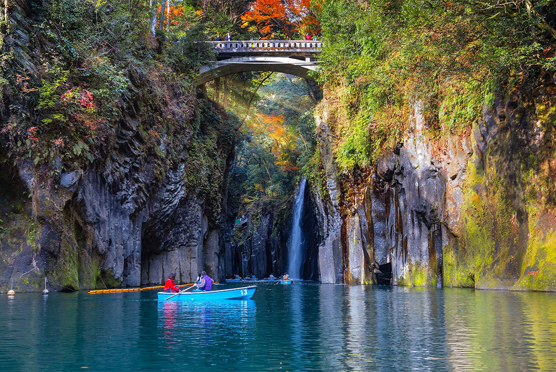 Travellers row boats through Takachiho gorge with waterfall and bridge as it turns to autumn