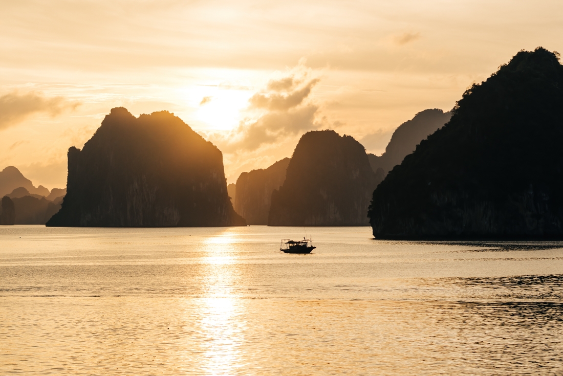 A small fishing boat crosses the waters of Ha Long Bay at sunset