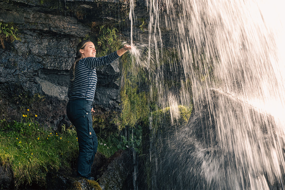Woman standing behind Gluggafoss waterfall with her hands in the streaming water