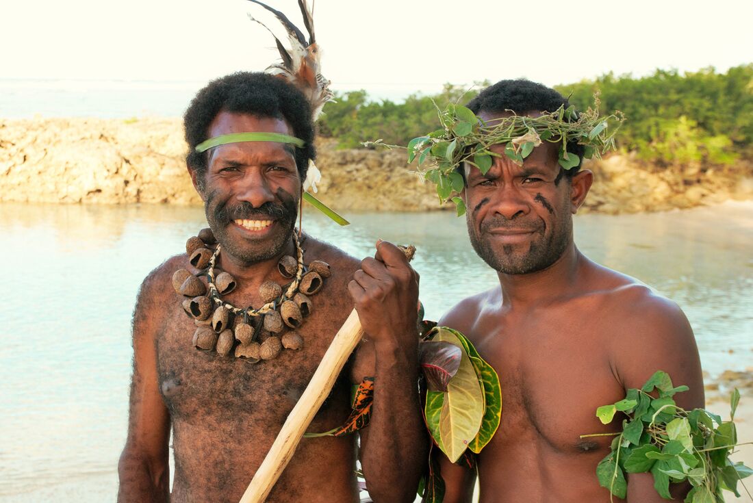 A medium shot of two smiling shirtless men. One is wearing a tree nut necklace and the other is wearing a crown made of leaves.