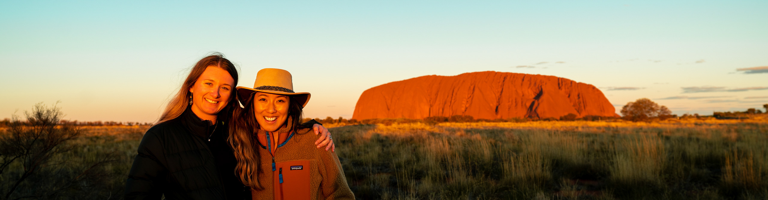 Two women stand in front of Uluru, having their picture taken during a bright red sunset 