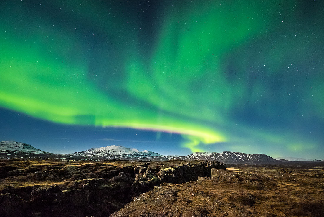 Green waves of the Northern Lights undulate over the rocky mountainous landscape of Thingvellir National Park, Iceland