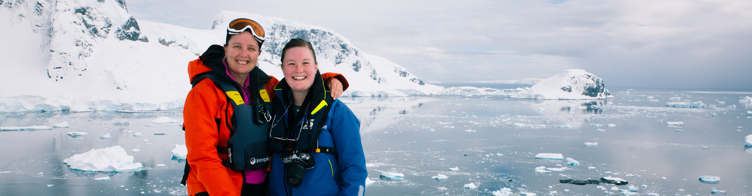 Pair of travellers smiling standing on an ice platform in the Antarctic Circle, with waters behind 
