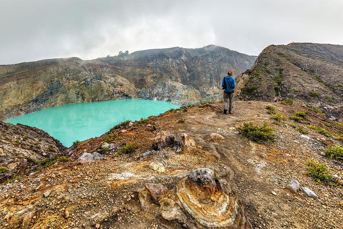 Traveller walks near cerulean blue acid lake at peak of Mount Kelimutu, Flores, Indonesia