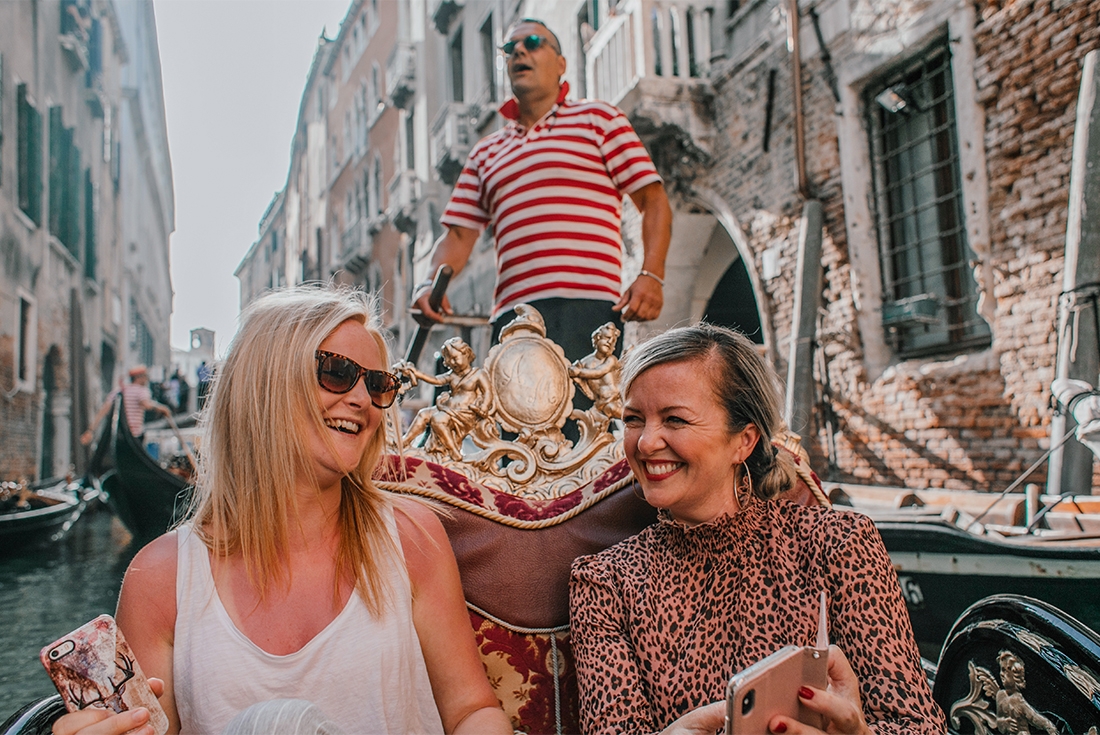Two travellers seated in a gondola in Venice while the gondolier navigates behind them