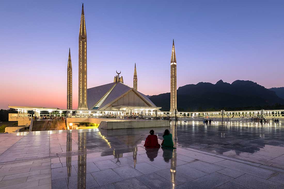 Two people sit and look at the Shah Faisal Mosque during purple sunset in Islamabad