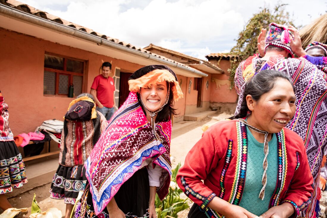 An Intrepid traveller wearing traditional Peruvian clothes smiles at the camera, as a local woman walks in the foreground and other people try on clothes in the background