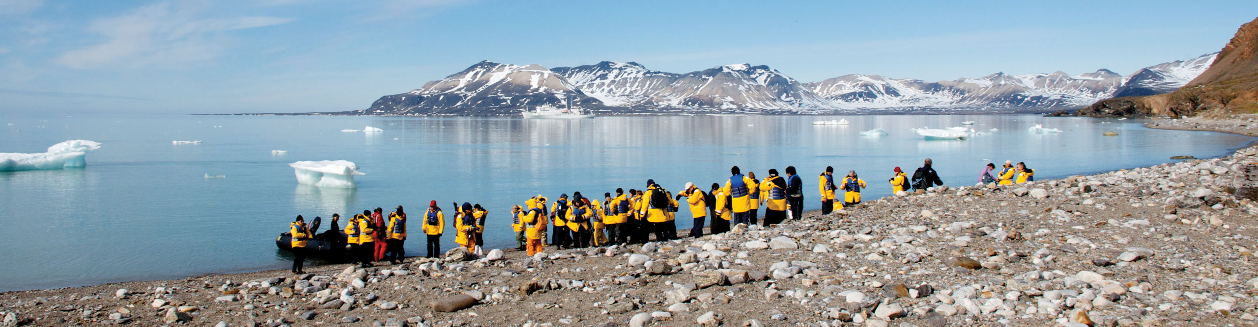 Group wearing yellow jackets on shoreline with icebergs in the distance on a sunny day in the Arctic