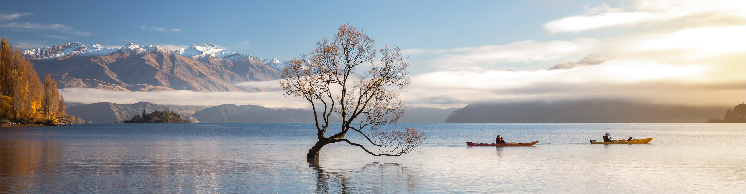 kayakers on the water by the famous tree in the water at Lake Wanaka with mountains in the distance