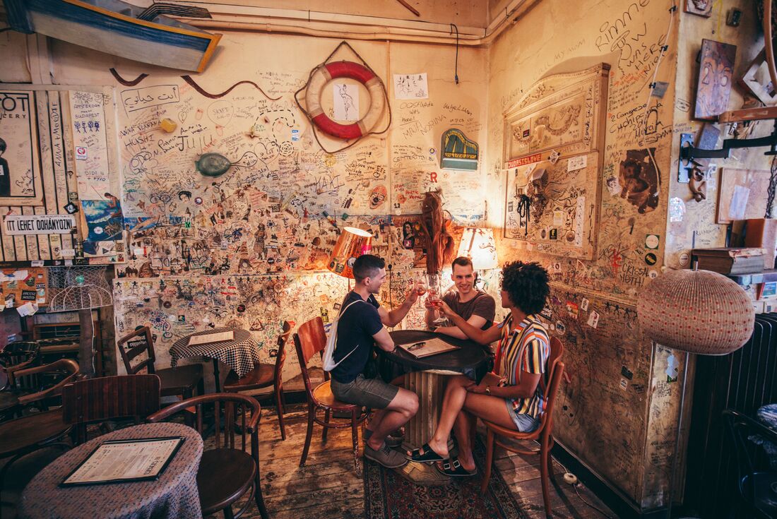Three young people toast their glasses as they sit in a heavily-graffitied dive bar