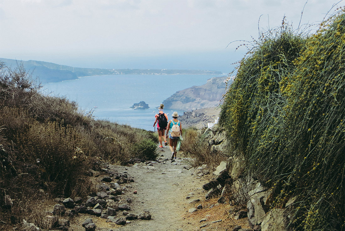 Two travellers on the caldera hiking track from Fira to Oia on Santorini with the Aegean Sea visible beyond