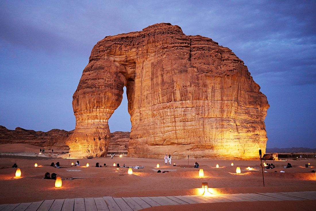Travellers seated in the sand enjoying twilight at Jabal Alfil, elephant rock, Al Ula, Saudi Arabia