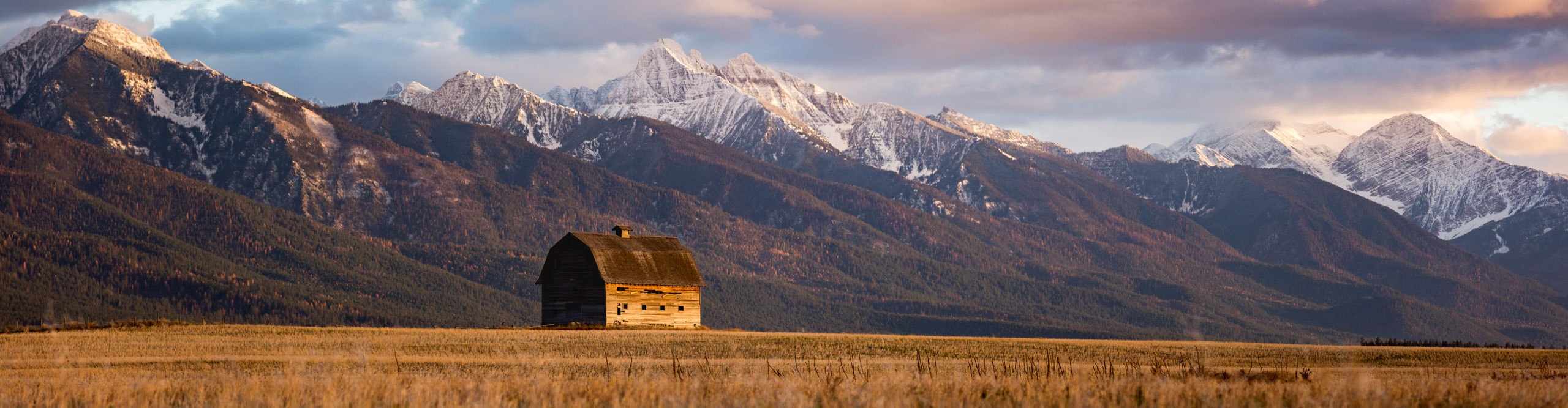 Barn in field with snow topped mountains in the distance, as the sun goes down, Pablo, Montana, USA