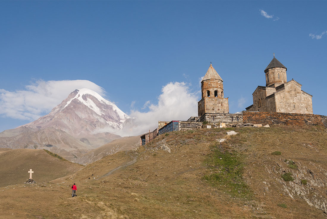 Georgia, Stepandsminda, Tsmina Sameba Church, 14th c, with Mt Kazbek and person in red approaching church on trail