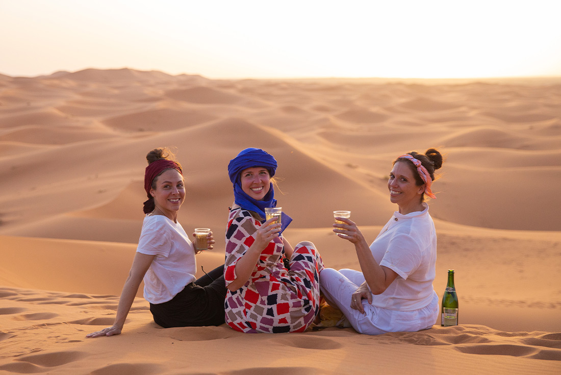 Group of female travellers enjoying a drink in the Sahara desert while watching the sunset in Morocco
