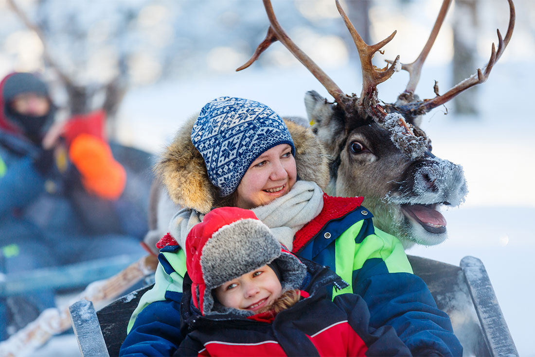 A friendly reindeer comes up behind a mother and child before a sleigh ride in Finland